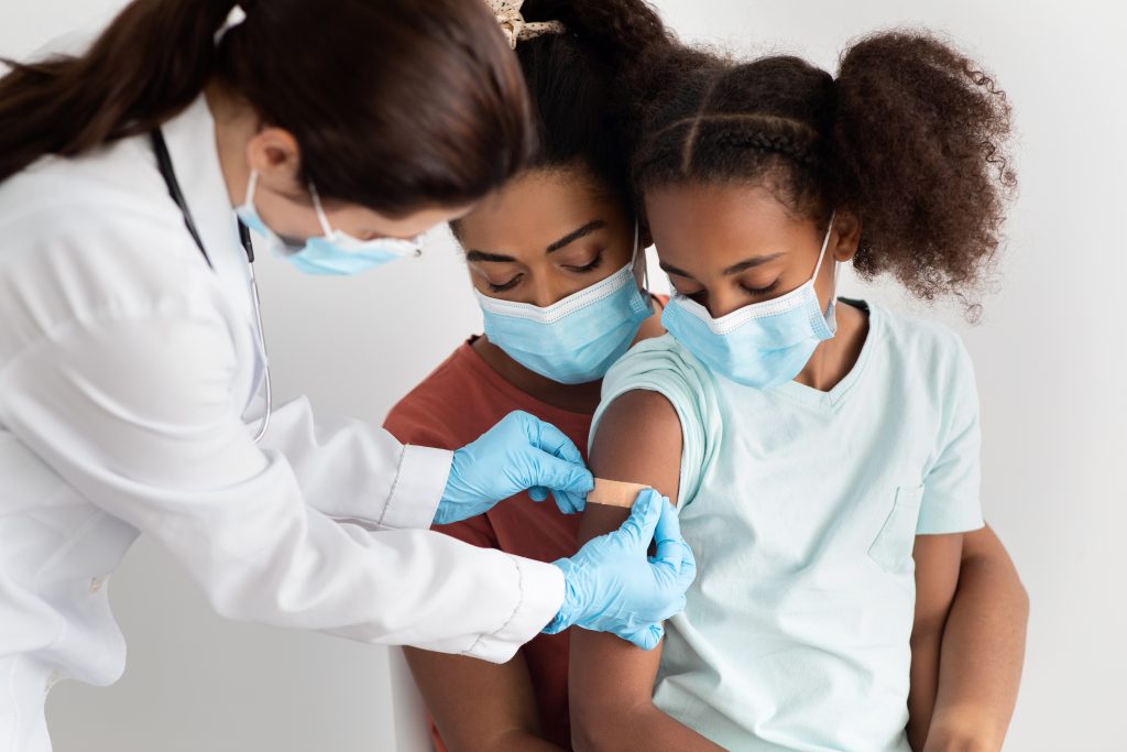 African American family mother and adorable daughter during vaccination at clinic, wearing protective face masks.  Doctor putting tape on baby's shoulder after coronavirus injection, close up