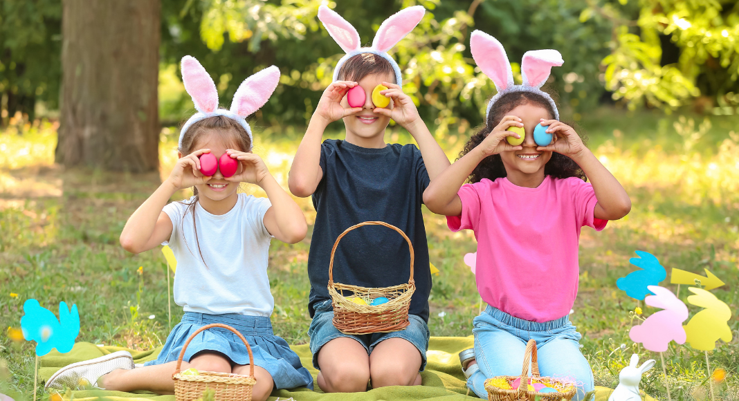 three children sitting with bunny ears headbands hold plastic eggs over their eyes